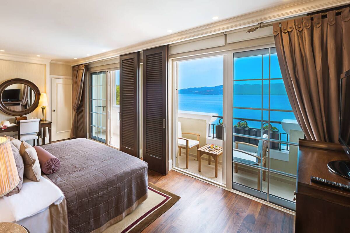 Un dormitorio amplio con una cama grande, una zona de estar con una mesa y sillas, televisión y un balcón con vistas al mar.