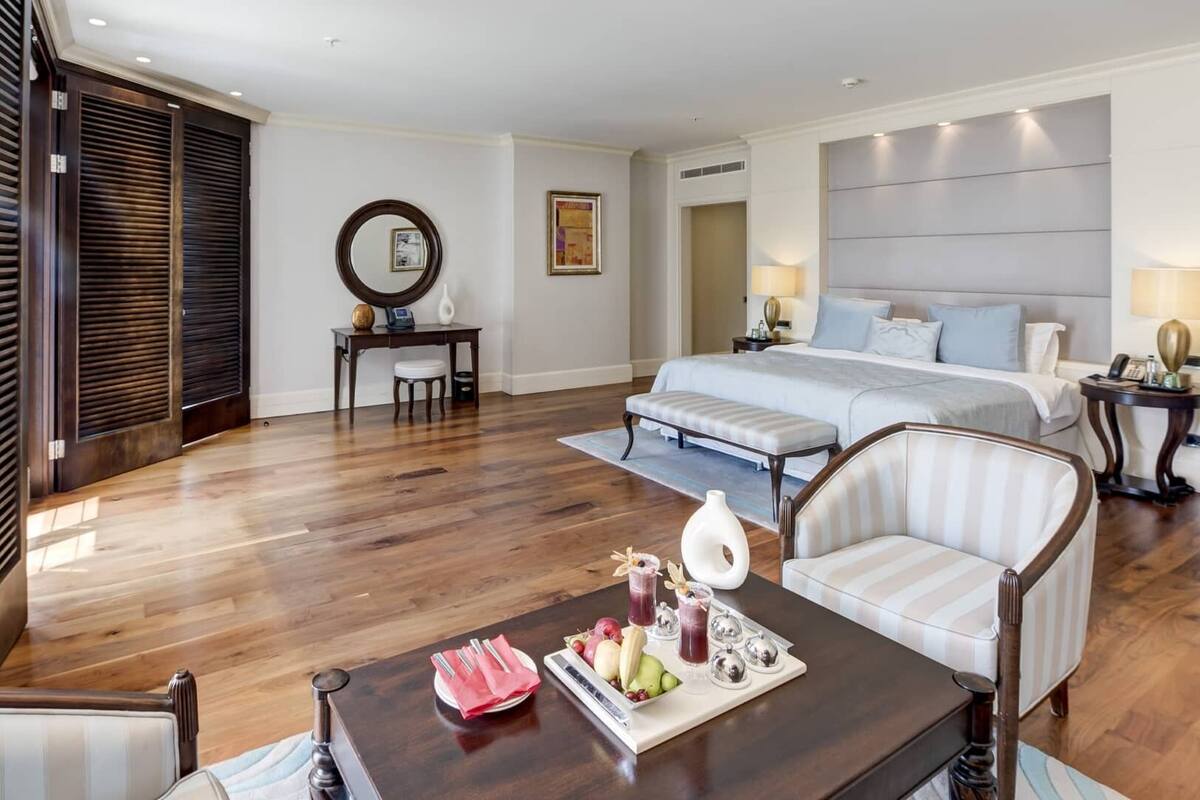 Habitación de hotel con una cama grande, un escritorio con silla, una mesa de comedor con una bandeja de fruta y un espejo redondo en la pared.