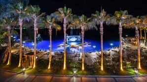 Vista noturna de um resort com área de piscina, palmeiras e palco.