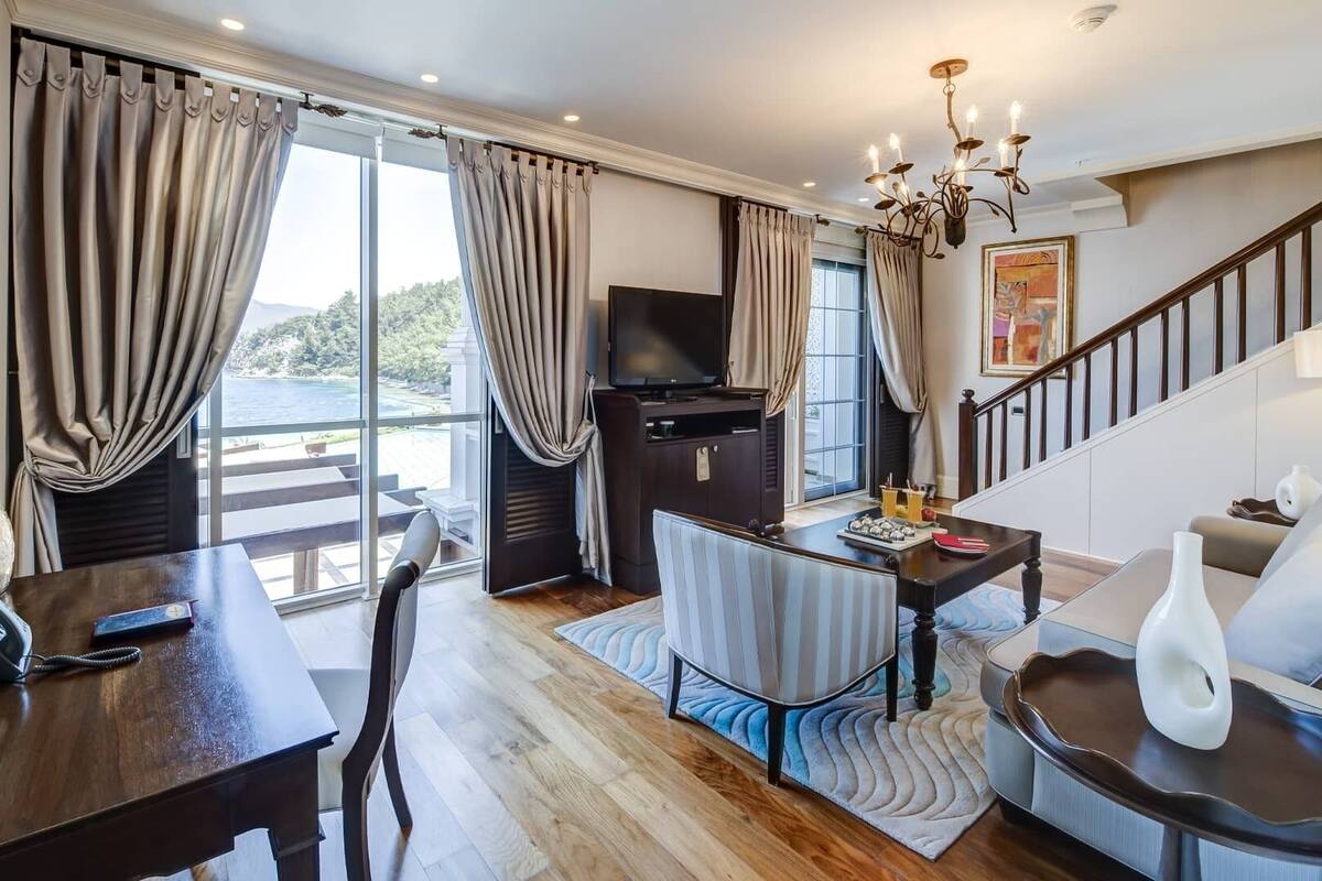 Amplia habitación con un gran ventanal que ofrece vistas al exterior, una mesa de comedor de madera, un sofá cómodo, un televisor y una escalera.