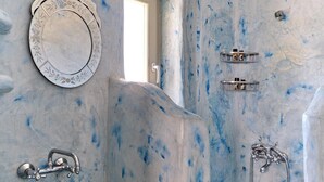 A bathroom with a marble-like wall design, a sink with a faucet, a shower with a handheld showerhead, and a mirror with a decorative frame. - Studio Eleftheria, Apleton Villas Paros (Paros)