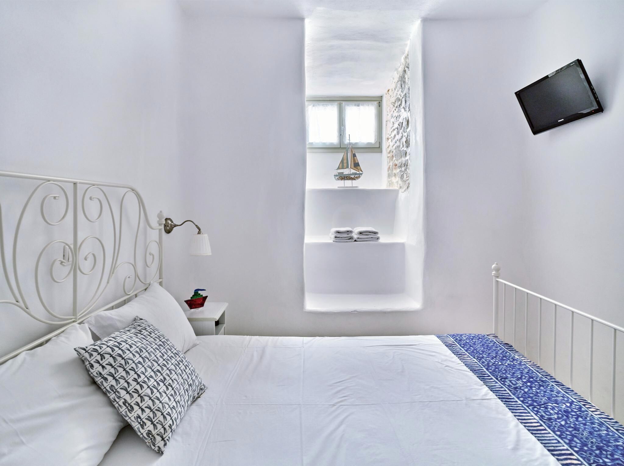 Une chambre avec un lit blanc, une table de chevet, une lampe, une étagère avec des objets décoratifs et un téléviseur fixé au mur.