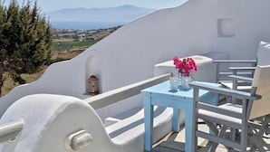 Habitación con paredes blancas y vistas al mar y a las montañas, una pequeña mesa con un florero y una silla.