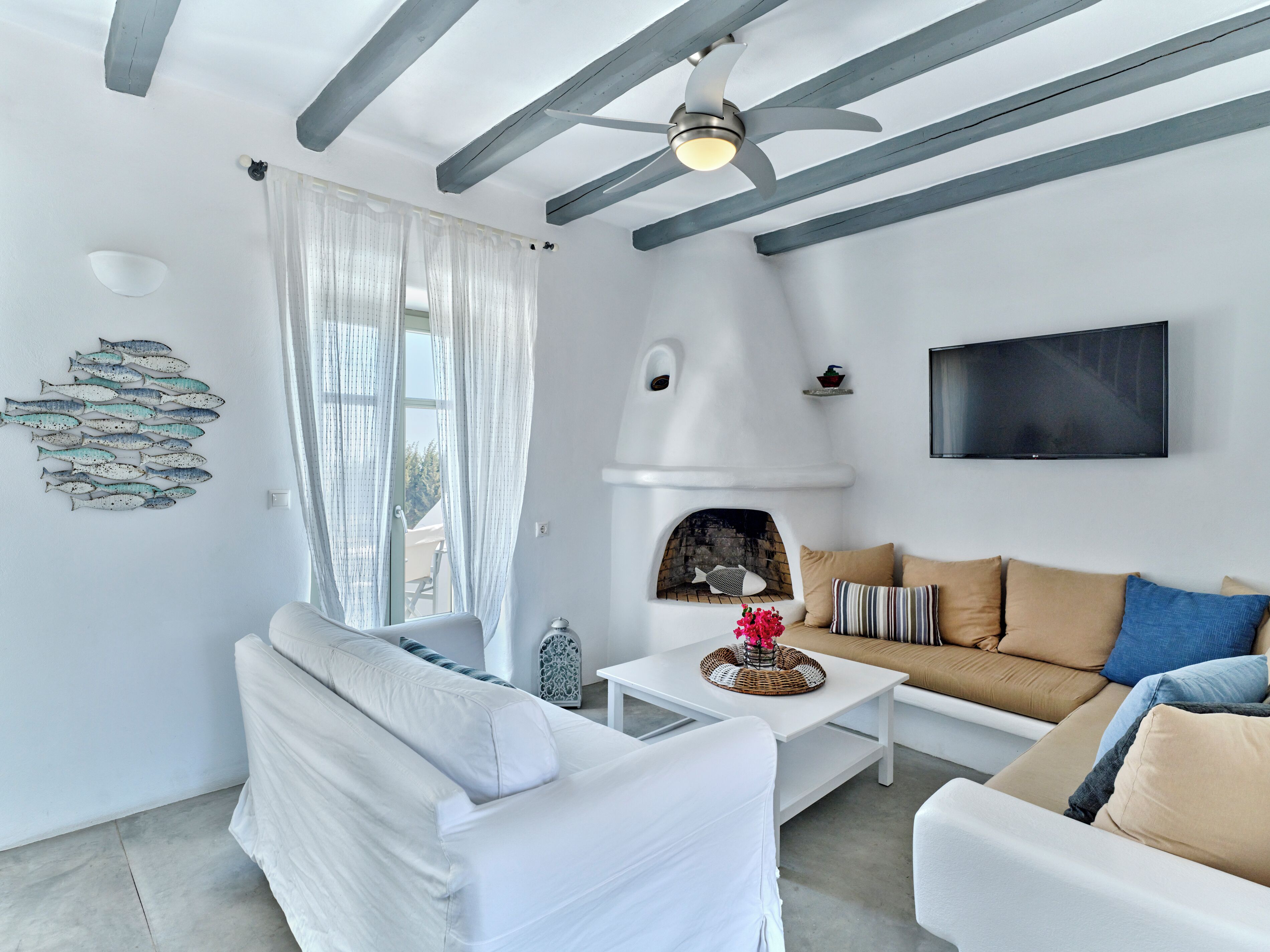 Un salon moderne avec un canapé blanc, une table basse blanche et un canapé beige. On y trouve une cheminée, un ventilateur de plafond et un téléviseur à écran plat fixé au mur.