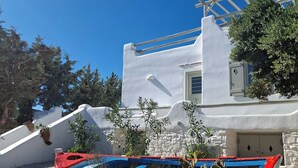 A boat with a red and blue hull, parked on a gravel surface with white stones, in front of a white building with a blue sky in the background. - Villa Pisti, Apleton Villas Paros (Paros)