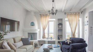 Un soggiorno con divano, poltrona e tavolino. È presente un caminetto, una TV fissata al muro e una vista sull'esterno attraverso una finestra.