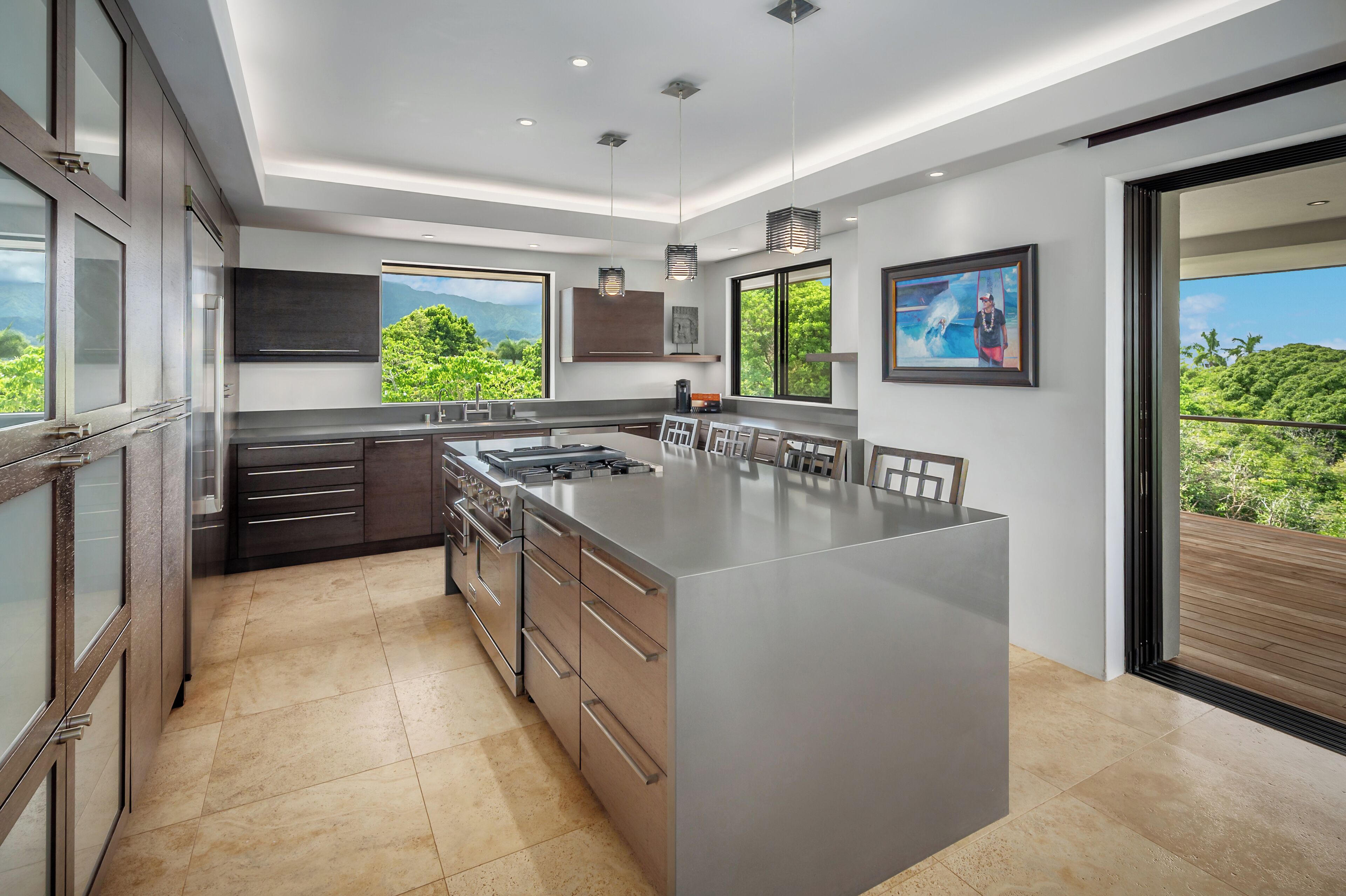 A modern kitchen with a large island, dark cabinetry, and large windows offering a view of greenery.