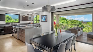 Een moderne keuken met een groot eiland, een eetgedeelte en een balkon met uitzicht op weelderige begroeiing.