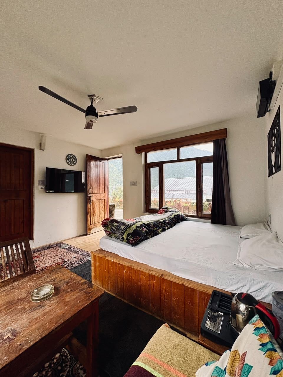 Ein Schlafzimmer mit Bett, Deckenventilator, Fernseher, Fenster mit Vorhängen und Blick ins Freie.
