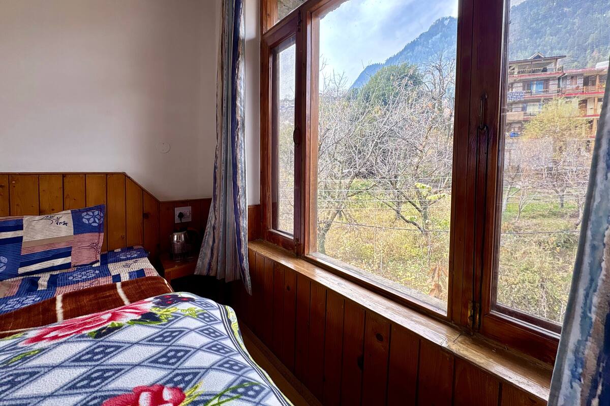 Un dormitorio con cabecera de madera, cama con colcha estampada, ventana con vista a árboles y edificios, y cortina.