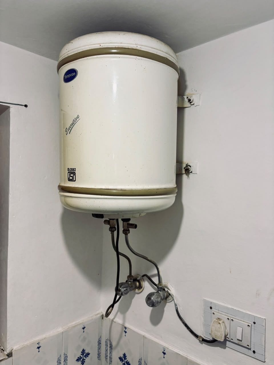 Een witte boiler aan een muur bevestigd, met zichtbare leidingen en een bedieningsschakelaar.