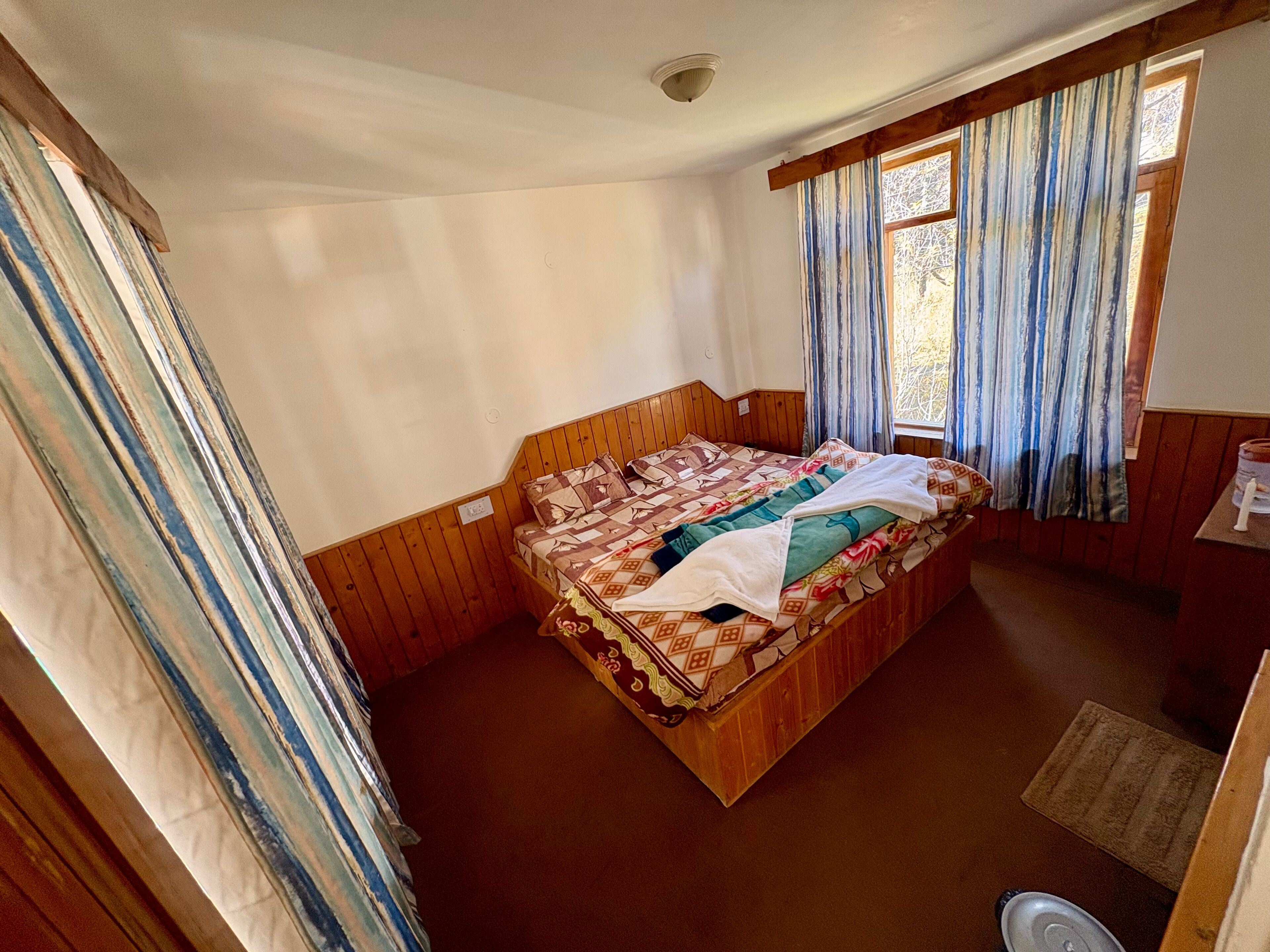 A room with wooden paneling, a bed with patterned bedding, a window with curtains, and a small table with a lamp.