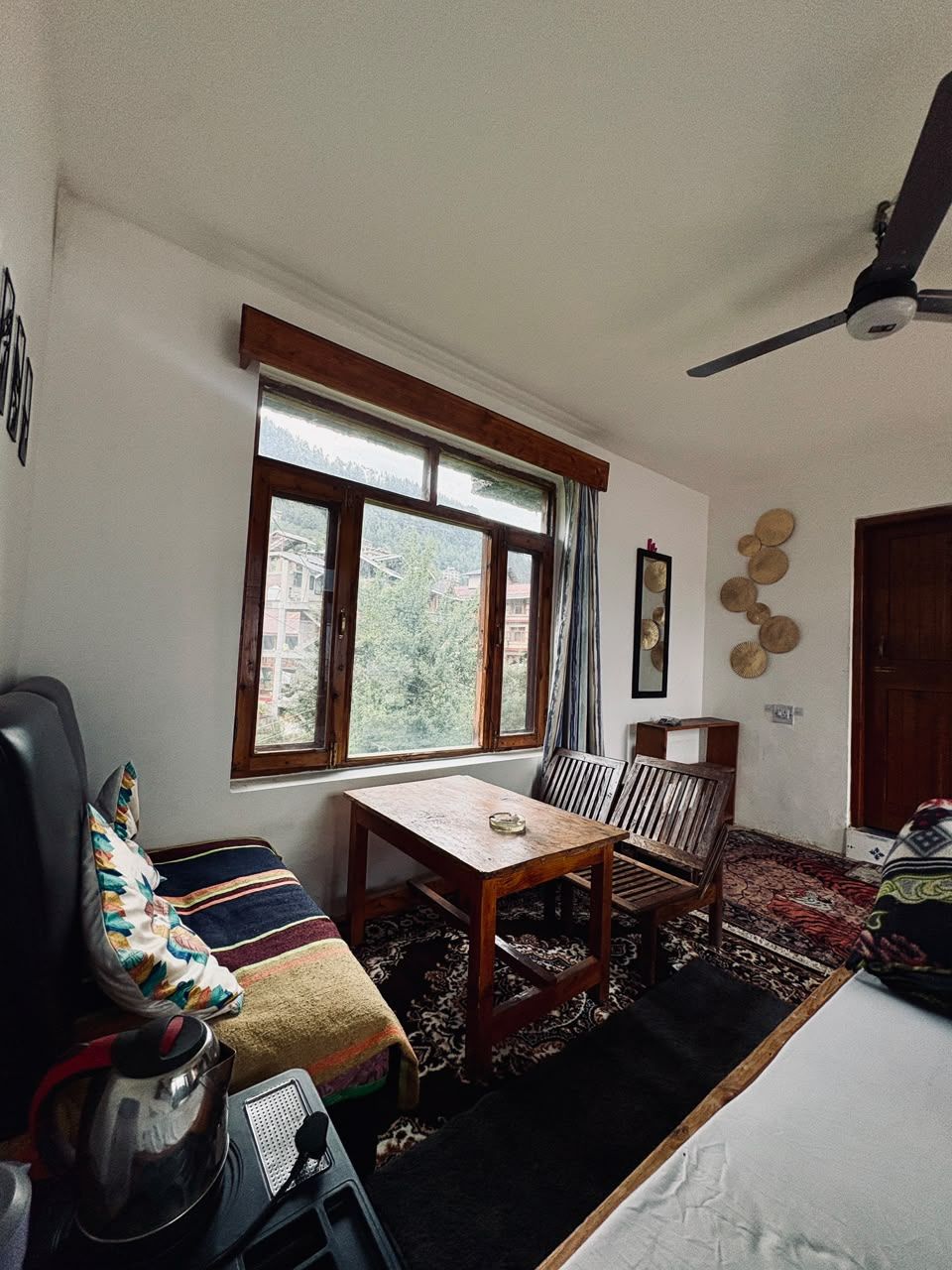 Una habitación con una mesa de madera, sillas, un sofá, un ventanal con cortinas, un ventilador de techo y vistas al verdor exterior.