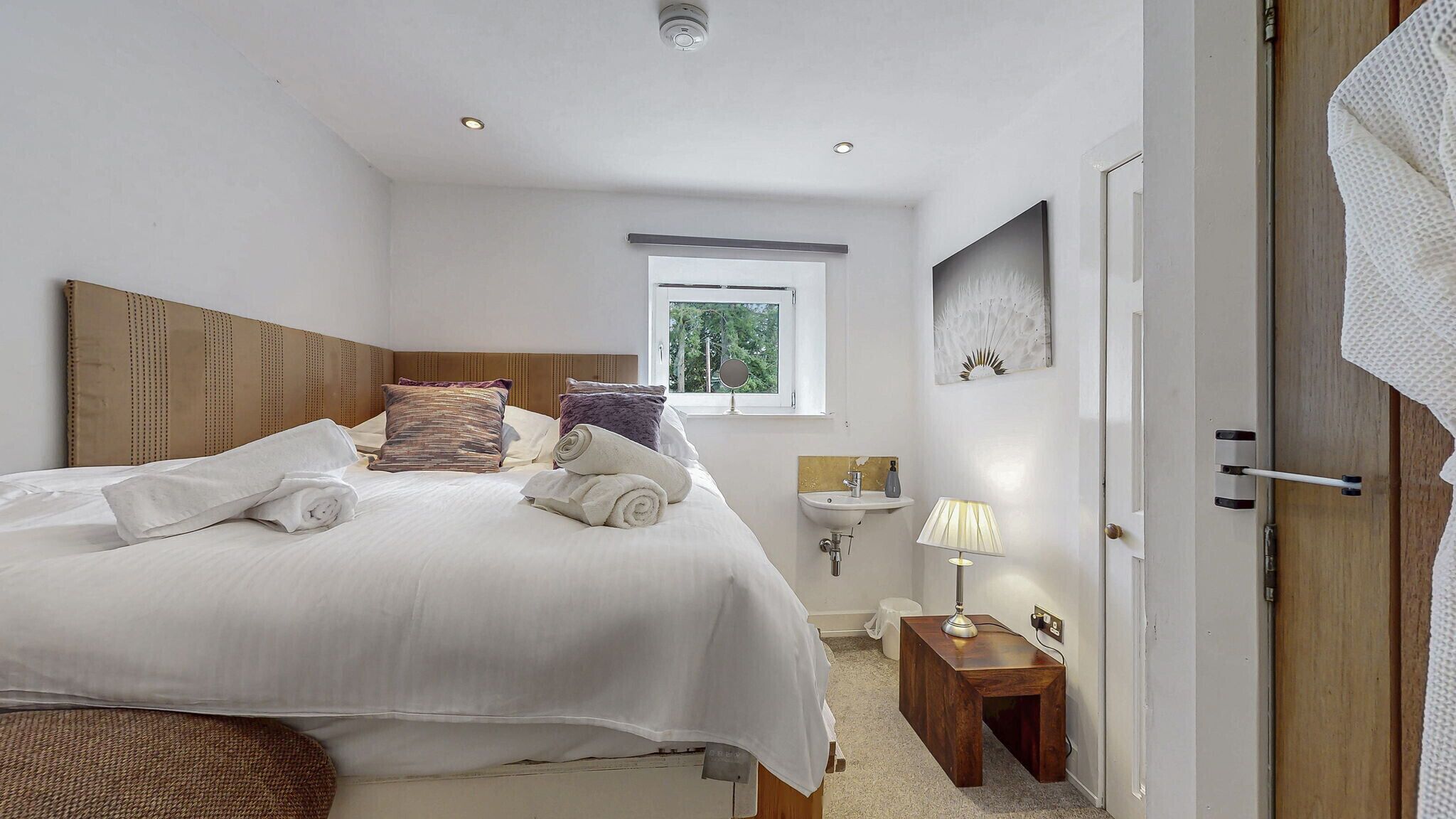 A neatly made bed with pillows and towels, a bedside table with a lamp, a window with a view of greenery, and a small bedside table with a vase.