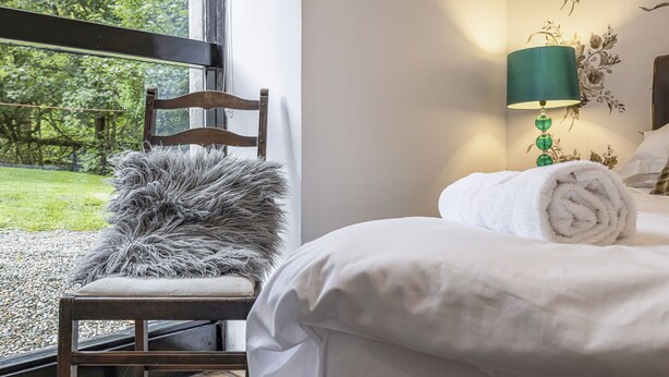 Ein Schlafzimmer mit einem Bett, einem Sessel mit einem flauschigen Kopfkissen, einer Lampe und einem Fenster mit Blick auf Grünflächen.