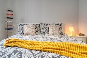 A neatly arranged bed with a patterned bedspread, two pillows with a floral design, a yellow throw blanket, a bedside lamp, and a bookcase with books.