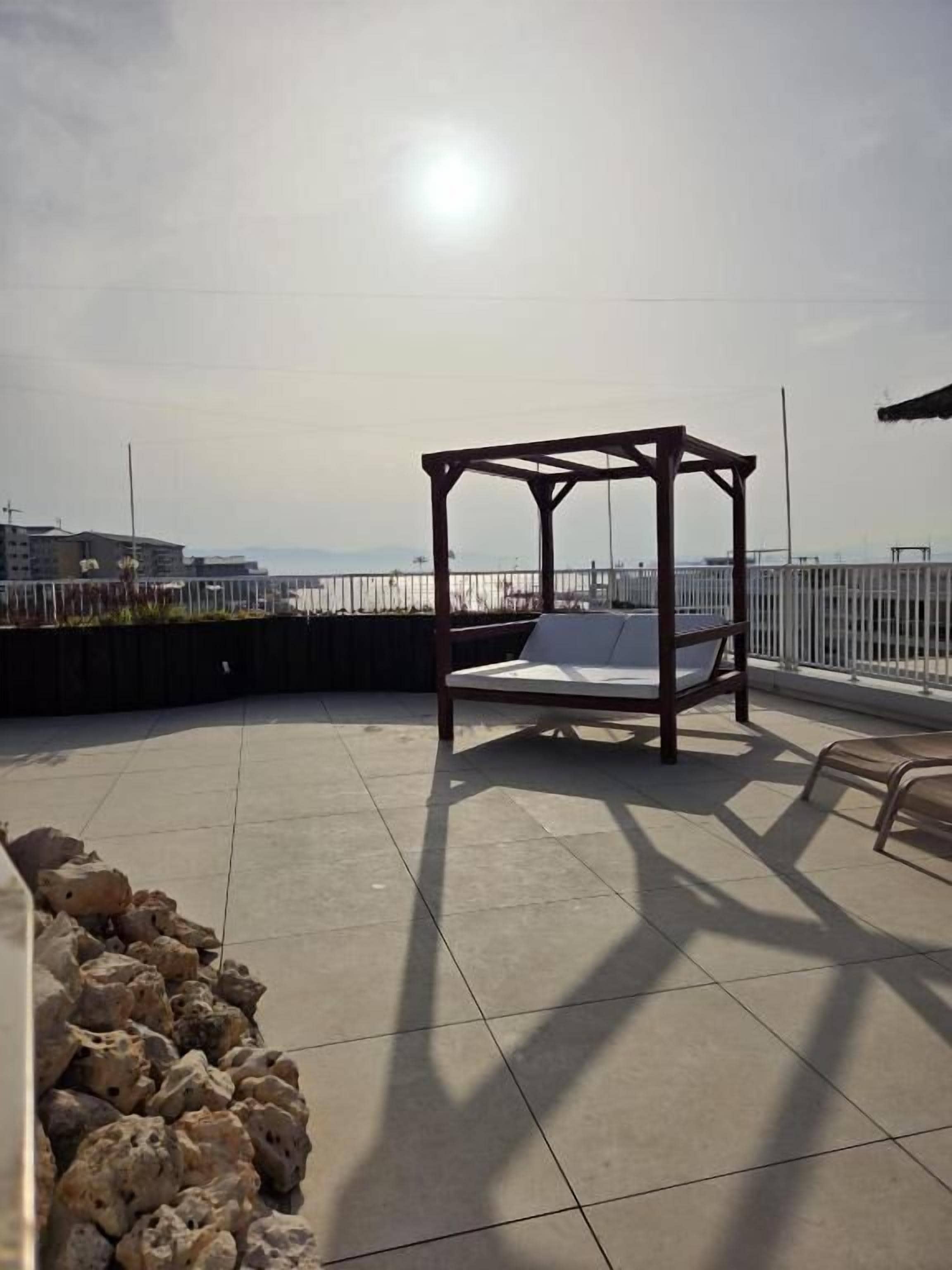 Un espacio al aire libre iluminado por el sol, con una pérgola de madera y un banco acolchado.