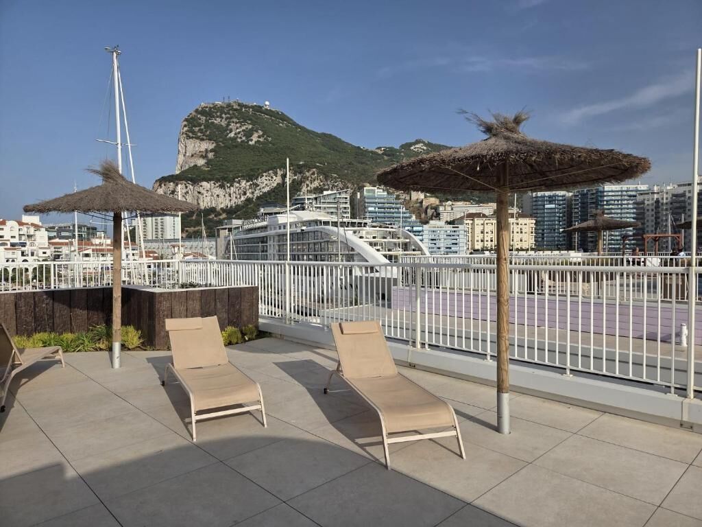 Terraço no telhado com duas cadeiras de lounge e um guarda-sol de palha, com vista para um porto e o horizonte urbano.