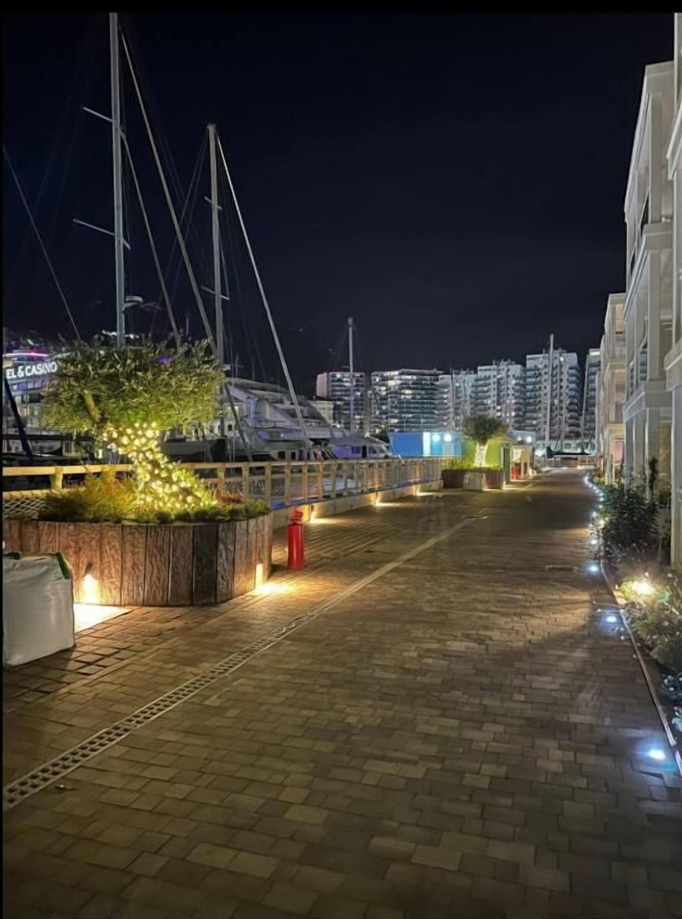 Marina nocturna con barcos, un muelle de madera y edificios al fondo.