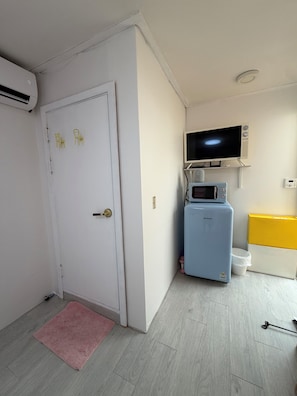 Une pièce de taille modeste avec une porte blanche, une petite kitchenette équipée d’un four à micro-ondes, une télévision et un réfrigérateur bleu.