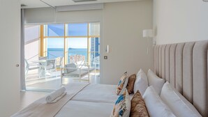 Een moderne hotelkamer met een groot bed, een balkon met uitzicht op zee en een zithoek met witte meubels.