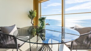 Een ronde glazen tafel met vier stoelen, een potplant en uitzicht op zee door grote ramen.