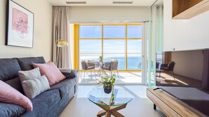Een moderne woonkamer met een grijze bank, een glazen salontafel, een flatscreen televisie en uitzicht op zee door grote ramen.