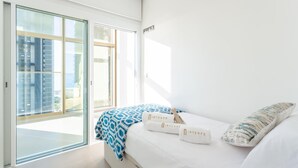 Ett modernt hotellrum med en stor skjutdörr i glas som leder ut till en balkong, en säng med vita sängkläder och två kuddar samt en blå, mönstrad pläd.