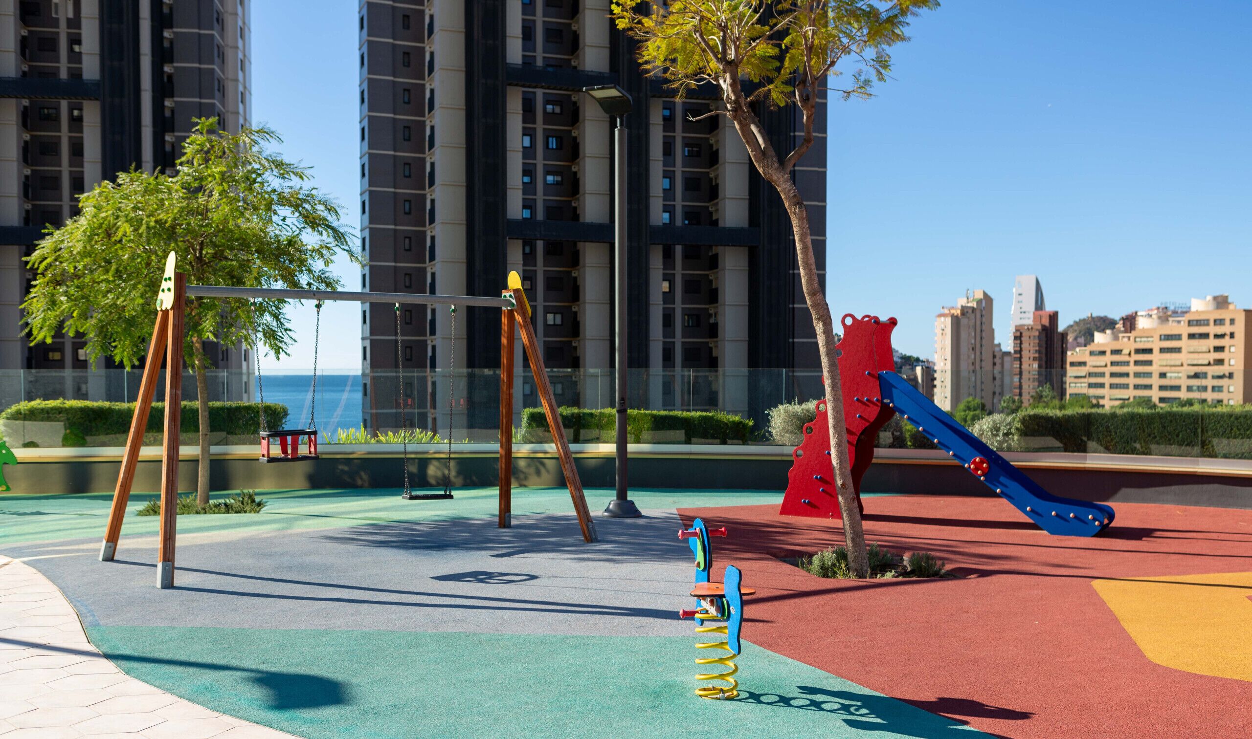 A playground with swings, a slide, and a climbing structure, surrounded by greenery and buildings.
