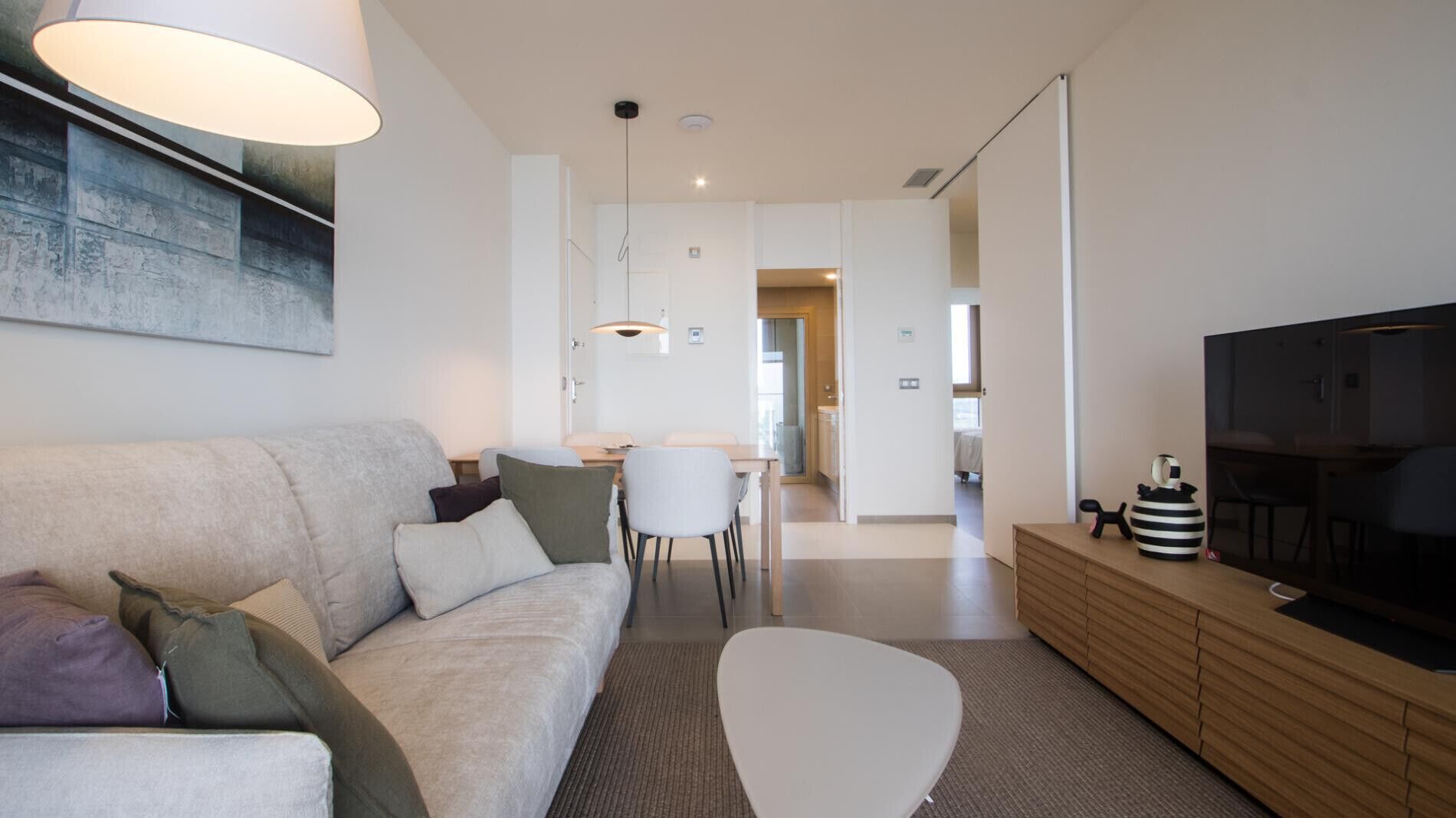 A modern living room with a sofa, a coffee table, a TV on a wooden cabinet, and a dining area in the background.