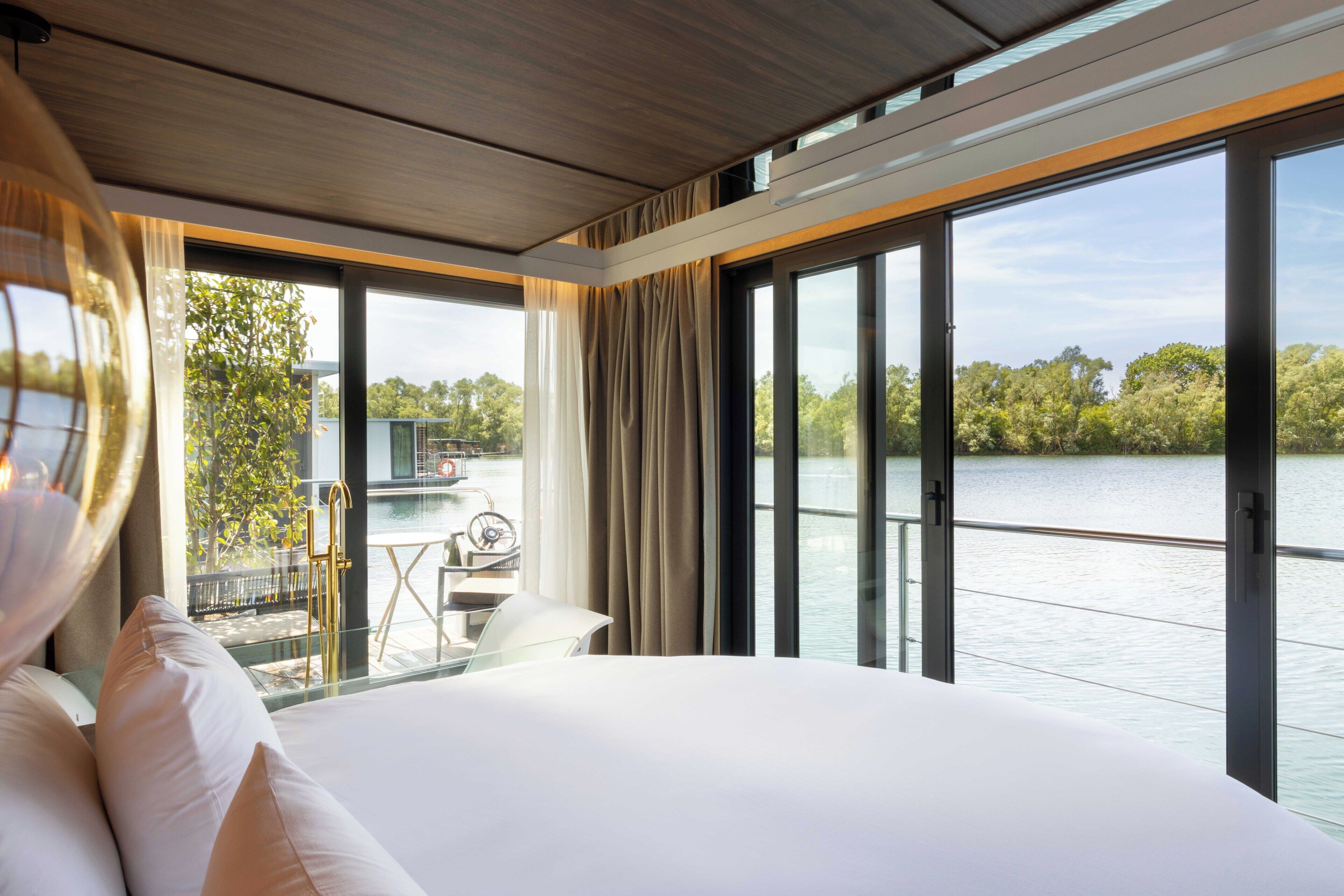Habitación de hotel moderna con una cama grande, vista a un lago y un área de comedor con mesa y sillas.
