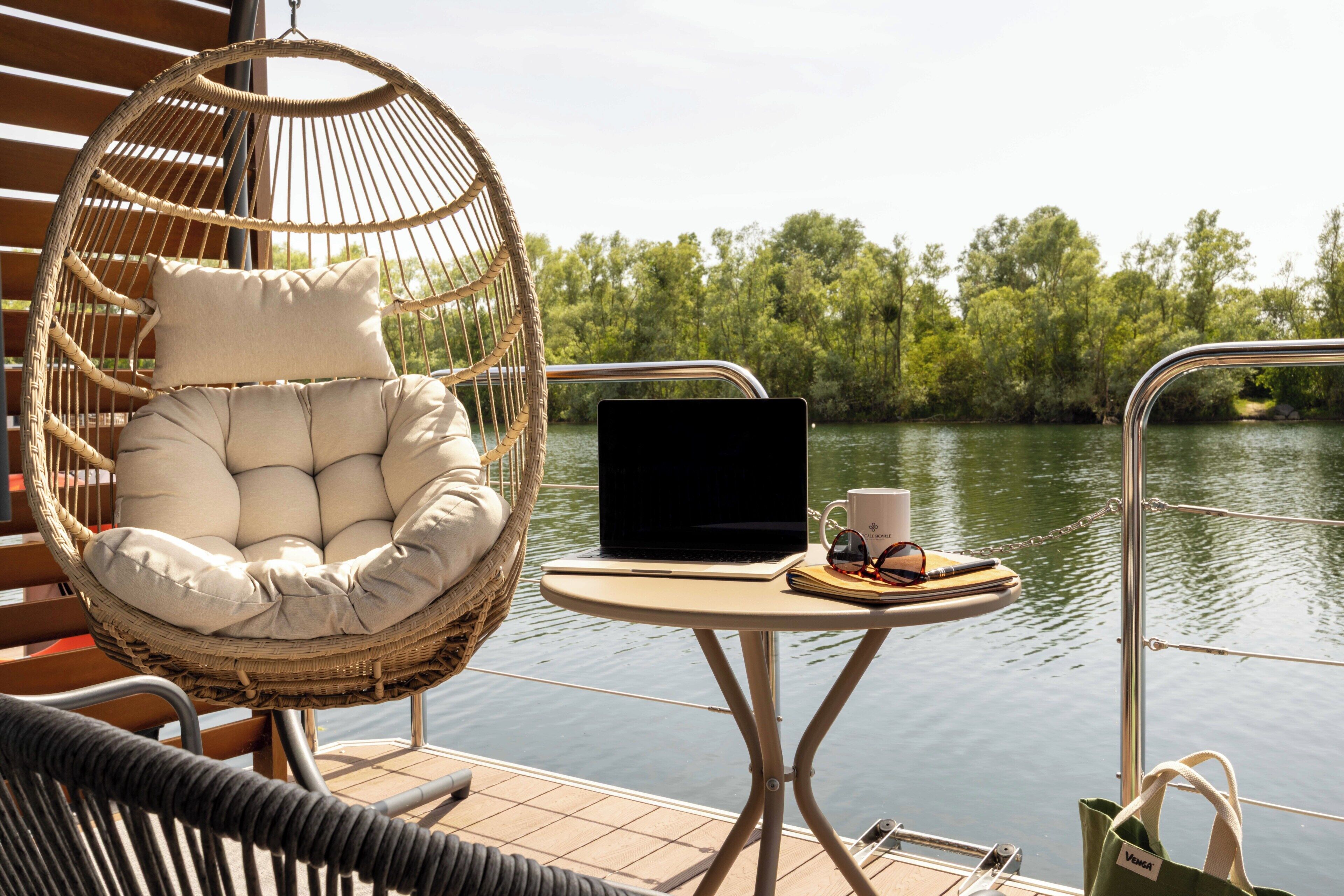 A wicker chair with cushions, a laptop, a mug, and sunglasses on a small table by a body of water.