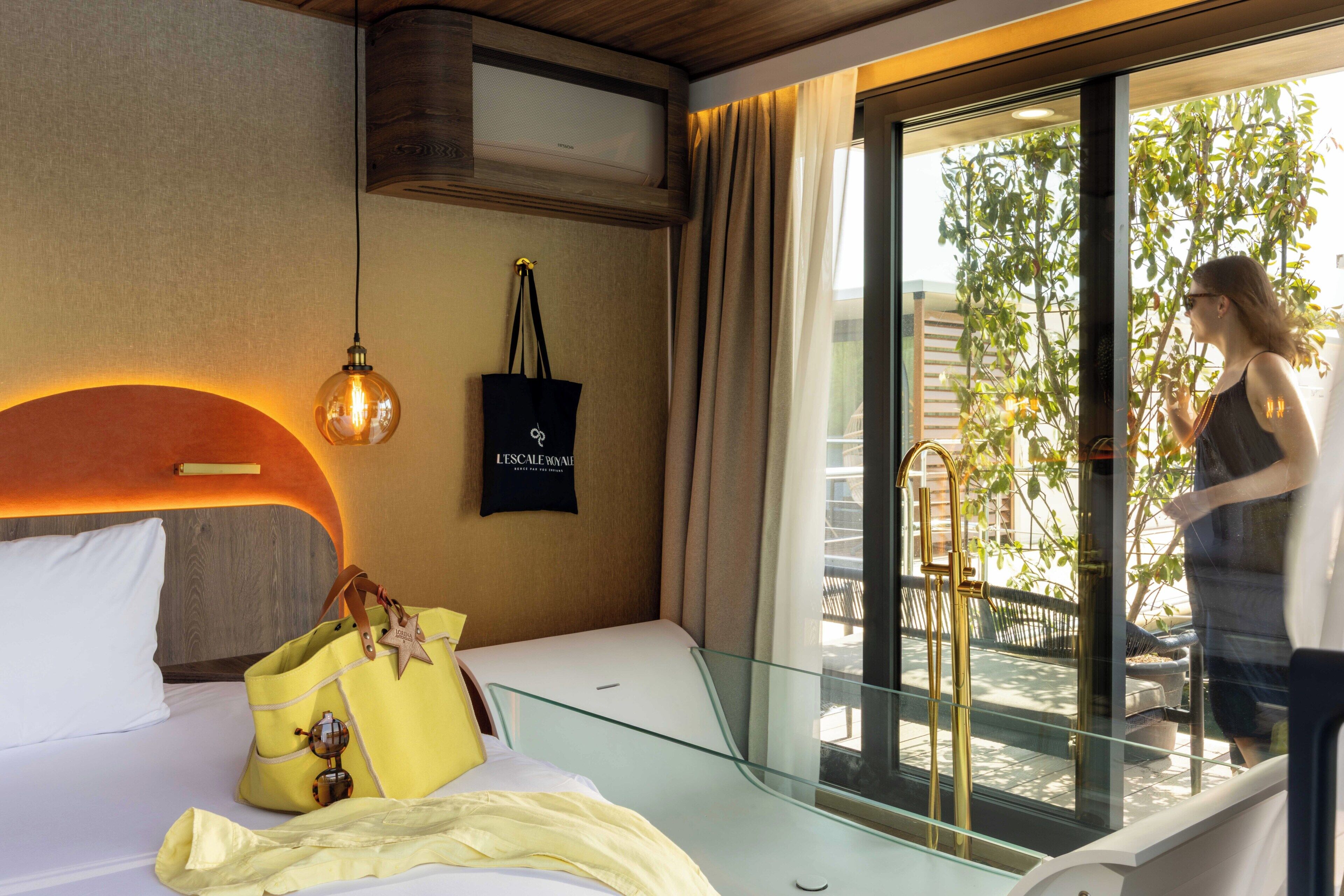 A hotel room with a bed, a hanging light fixture, a black tote bag, and a window with a view of greenery.