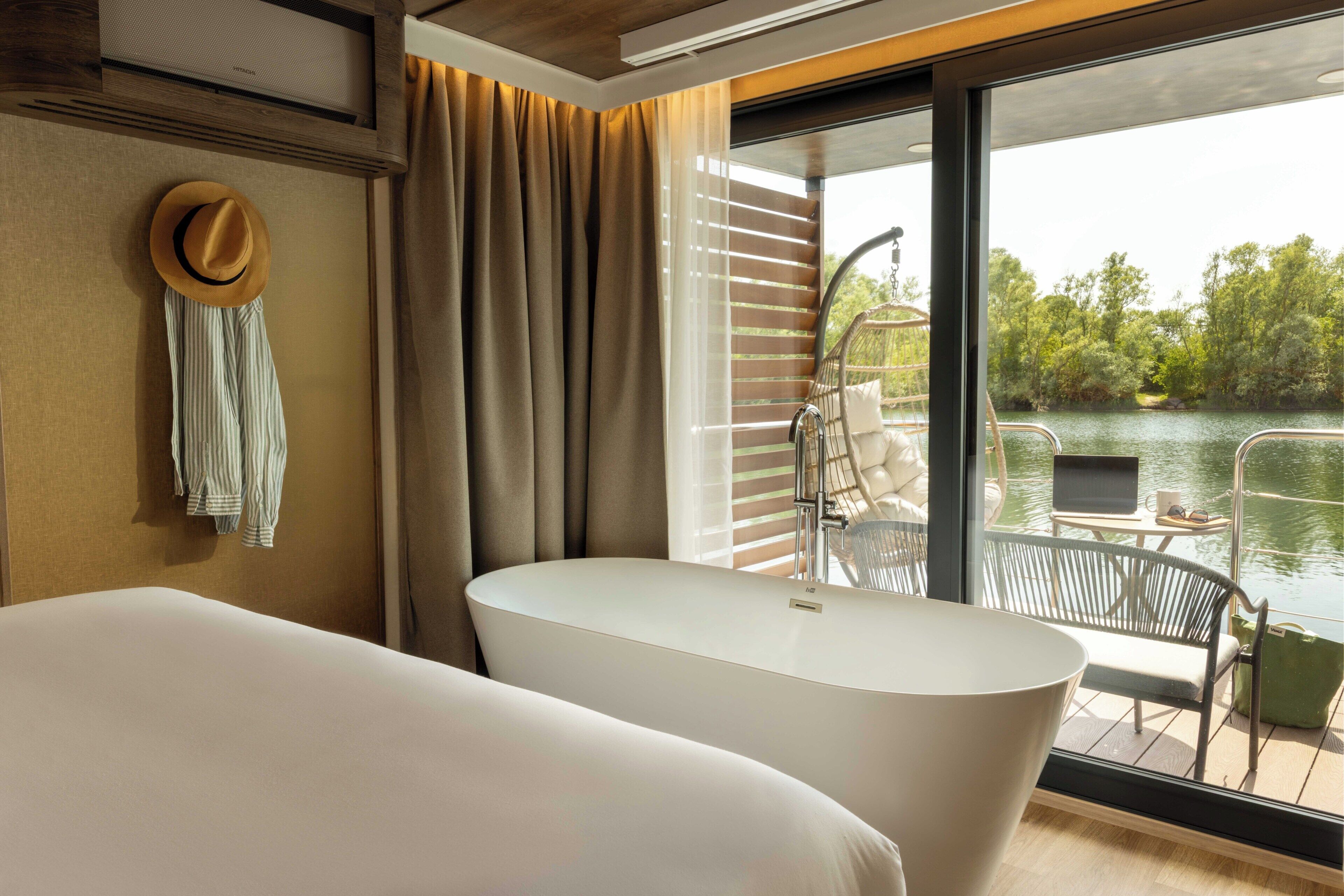 A modern bedroom with a large bathtub, a hanging hat, and a view of a lake through large windows.