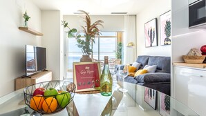 Un salon moderne avec un téléviseur à écran plat, une table à manger ornée de fruits et d’une bouteille de vin, un canapé avec des coussins et une grande fenêtre offrant une vue panoramique.