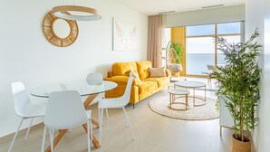 Een moderne woonkamer met een gele bank, witte stoelen, een glazen eettafel en een groot raam met uitzicht.