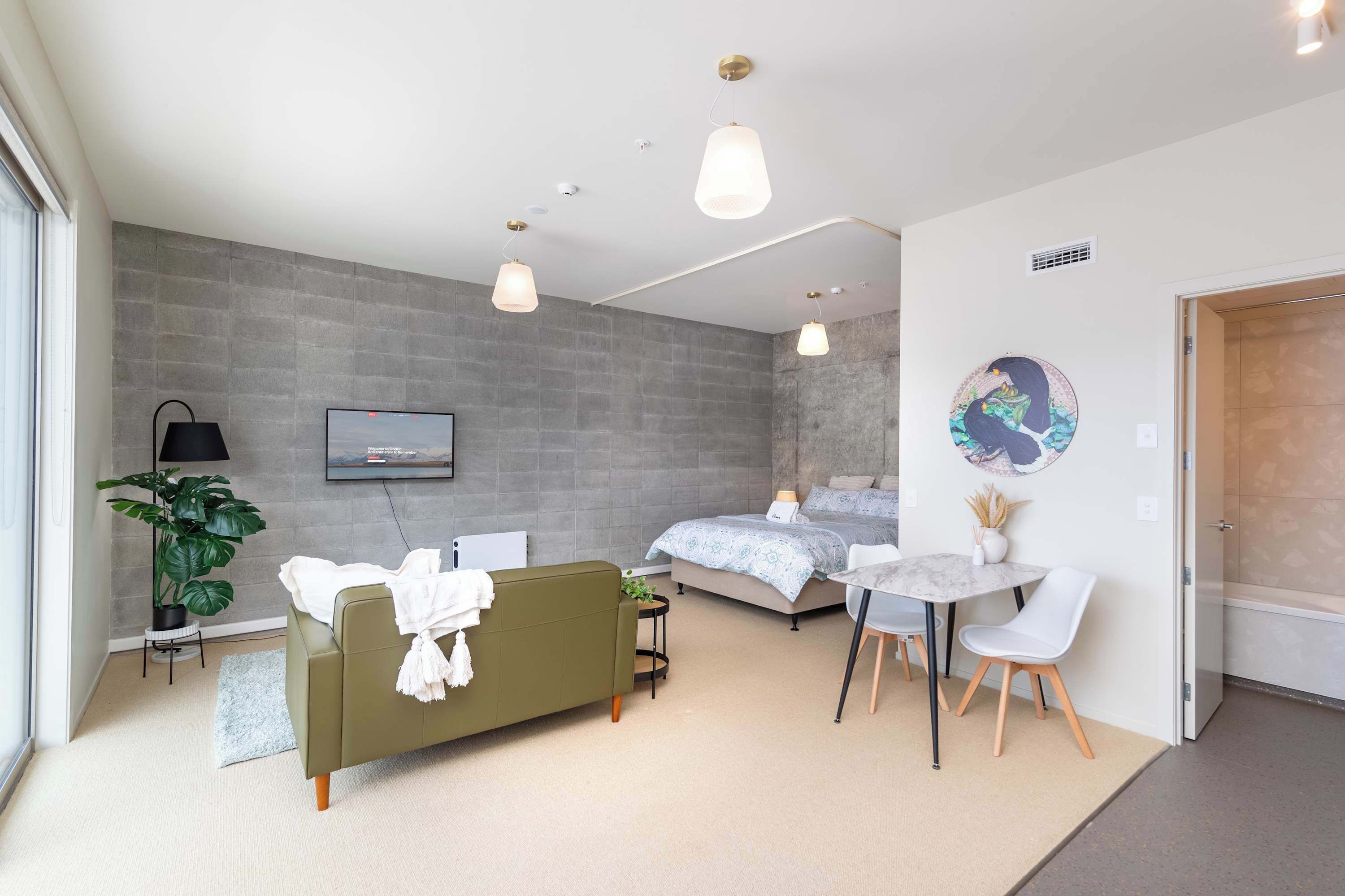 A modern bedroom with a bed, a sofa, a desk with chairs, a TV, and a plant.