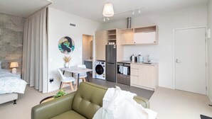Una camera d'albergo moderna con un divano verde, un'area pranzo con tavolo e sedie, una cucina con frigorifero e lavatrice, e un bagno con doccia.