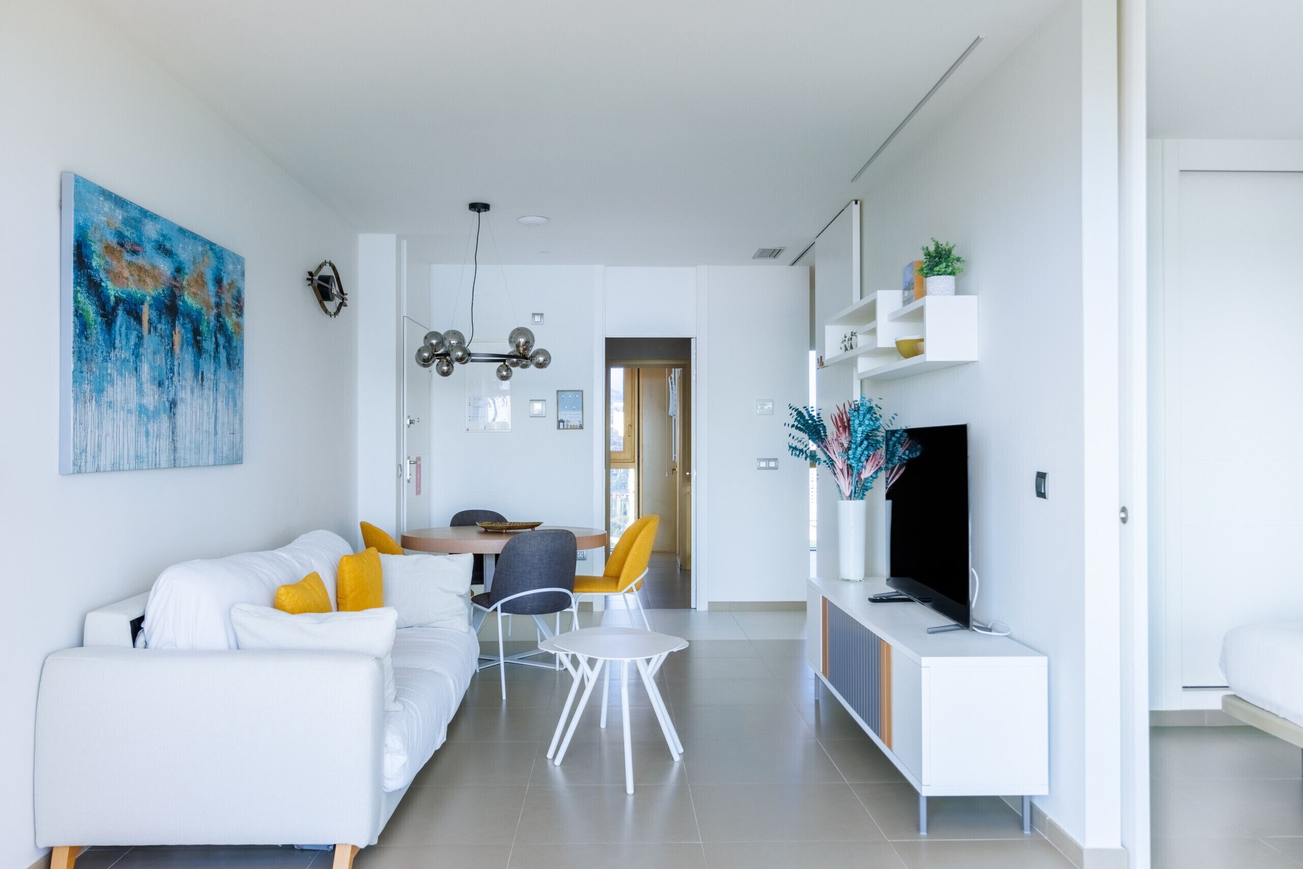 A modern living room with a white sofa, a dining table with chairs, a TV on a white cabinet, and a painting on the wall.