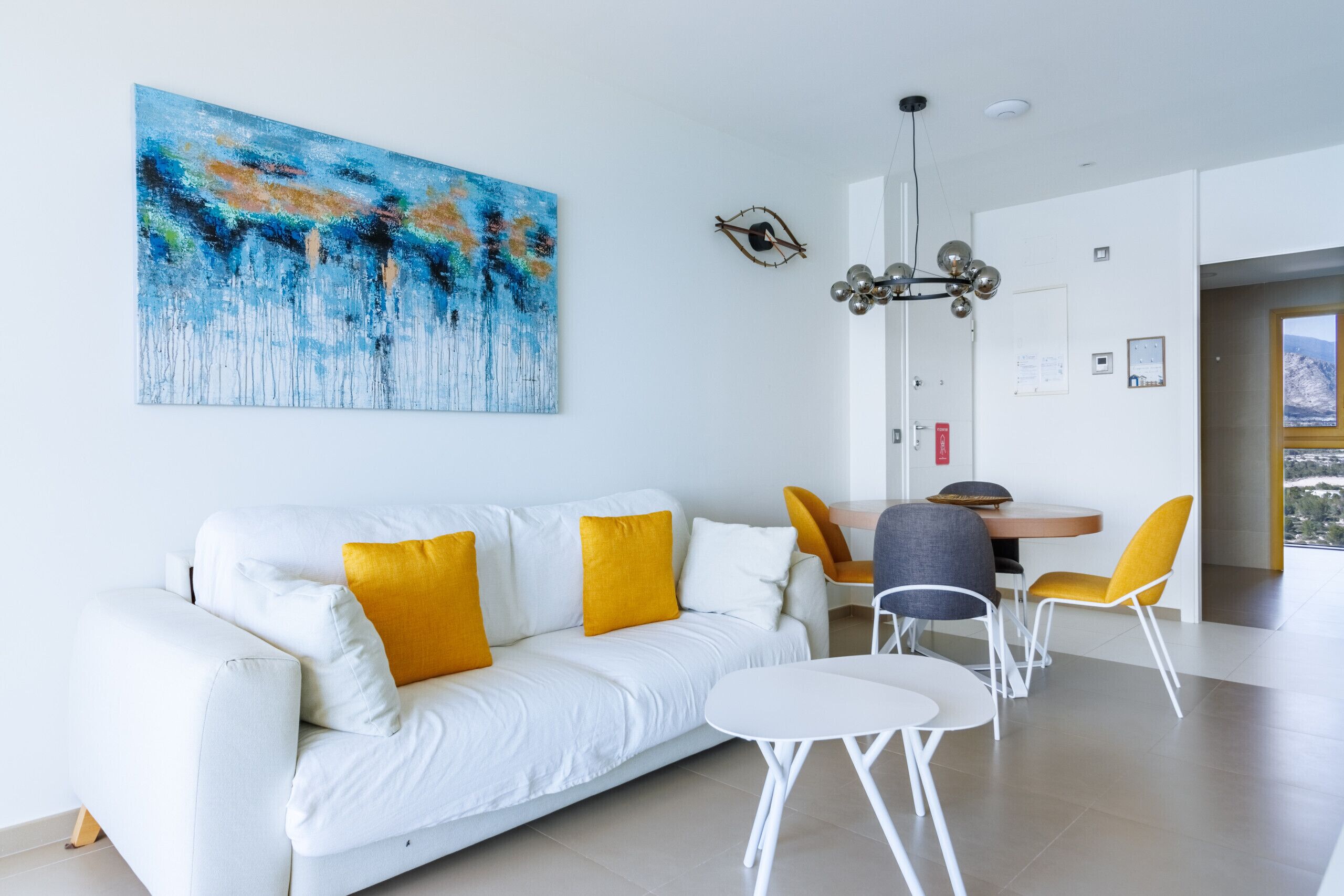 A modern living room with a white sofa, yellow cushions, and a large abstract painting. There is a dining area with a wooden table and yellow chairs.