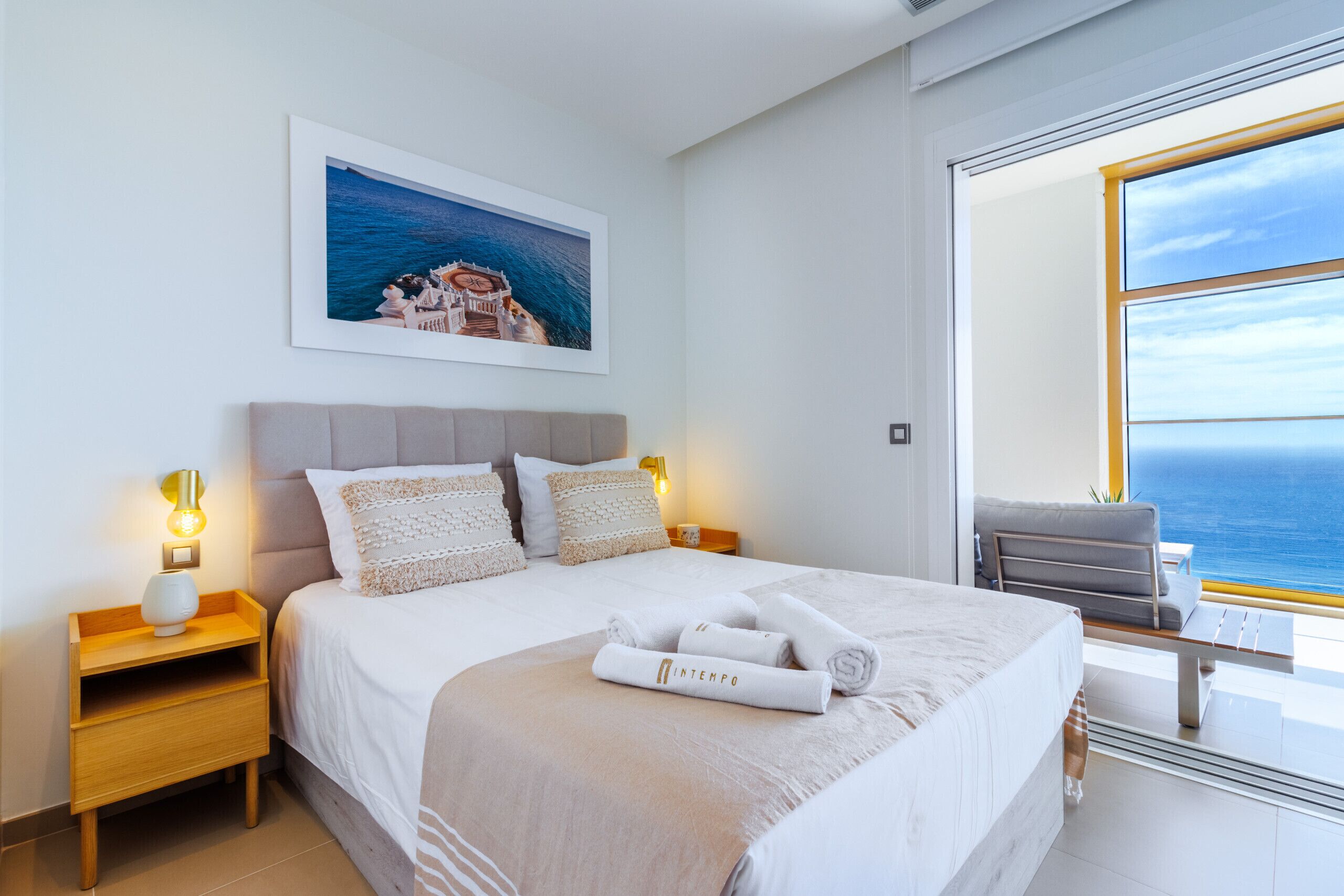 A modern hotel room with a large bed, a nightstand with a lamp, a framed picture of a coastal scene, and a view of the sea through a large window.