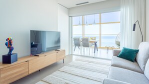 Una sala de estar moderna con una televisión de pantalla plana sobre un mueble de madera, un sofá gris con un cojín azul turquesa, y vista al mar a través de amplios ventanales.