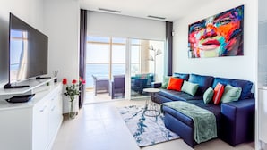 Una sala de estar moderna con un televisor de pantalla plana grande, un sofá con cojines coloridos, una mesa de centro de vidrio y vistas al océano a través de amplias puertas correderas.