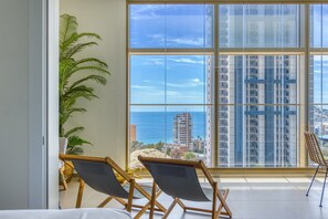 A room with a large window, two chairs, a potted plant, and a view of the ocean and buildings. - Intempo by Holuxe - 036 - 3rd floor (Benidorm)