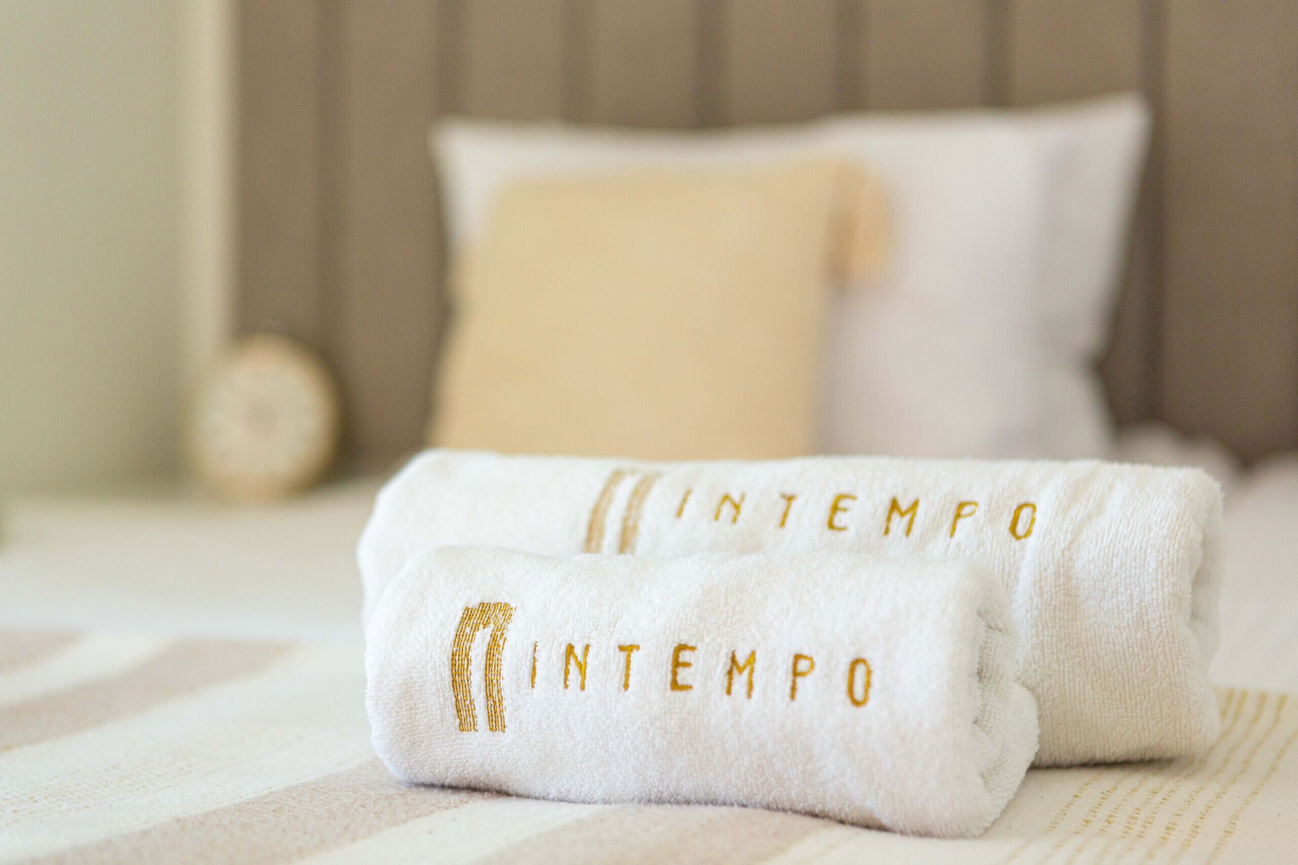 Two white towels with 'INTEMPO' embroidered in gold, placed on a bed with a striped bedspread.