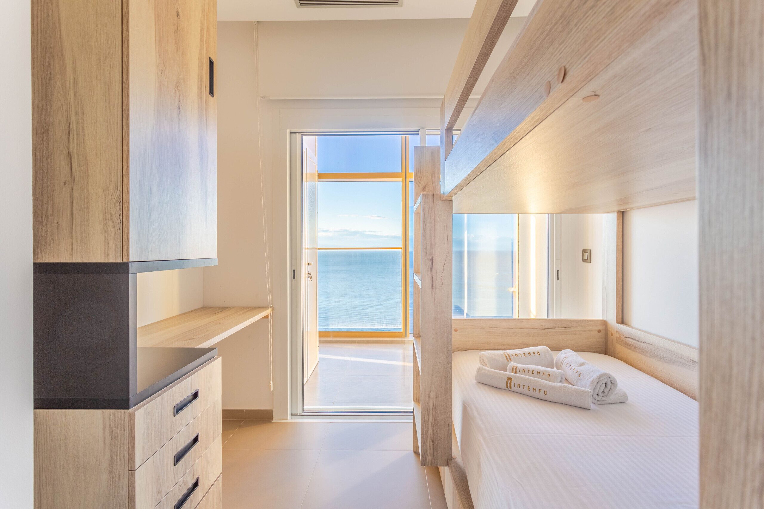 Habitació d'hotel moderna amb interior de fusta, un llit amb edredó blanc i vista al mar a través d'una porta oberta.