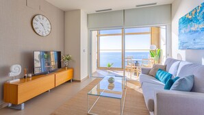 Una sala de estar moderna con un sofá, una mesa de centro de vidrio, un televisor sobre un mueble de madera y un amplio ventanal con vistas al mar.