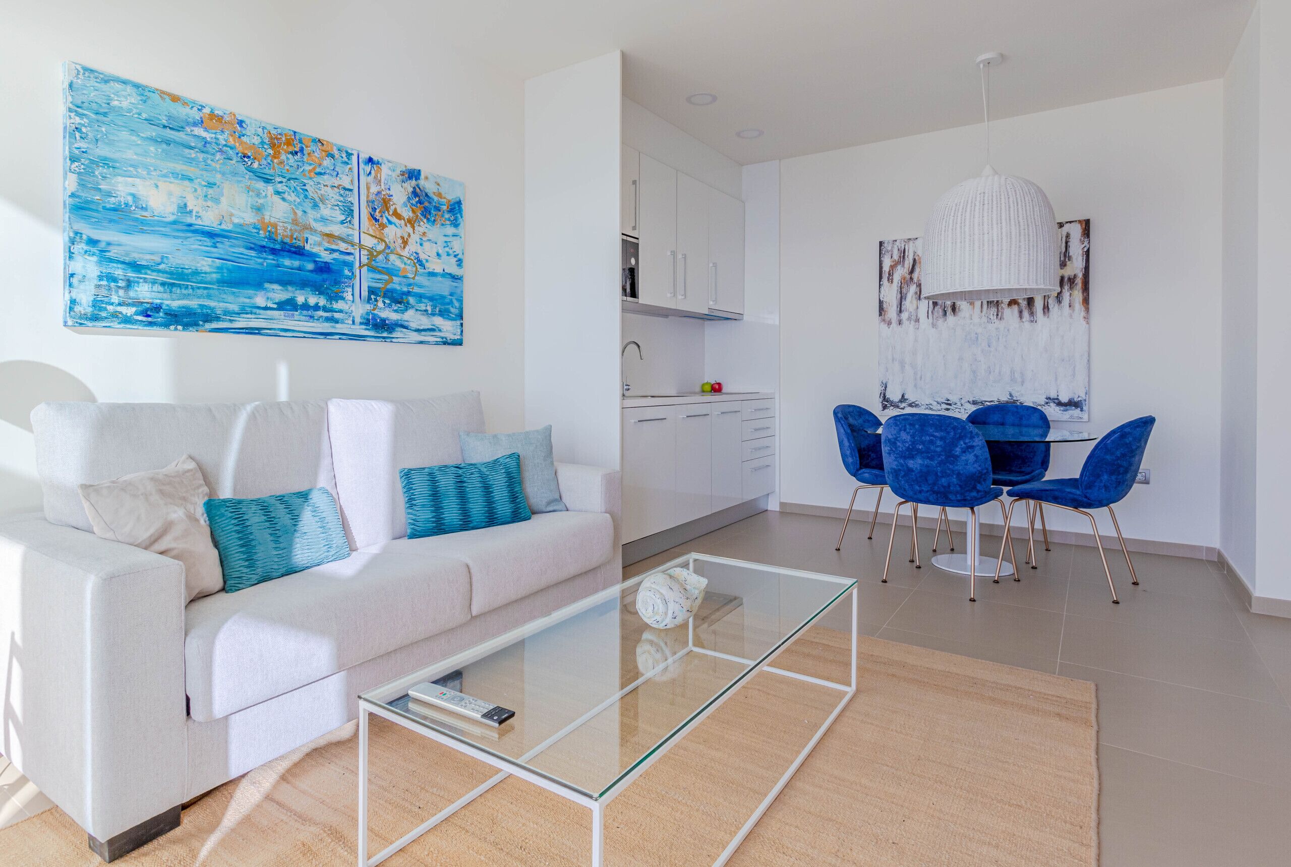 A modern living room with a glass coffee table, a white sofa with blue cushions, a dining area with blue chairs, and a kitchen with white cabinets.