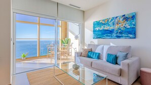 Una sala de estar moderna con una mesa de centro de vidrio, un sofá con cojines azules y un cuadro grande de un paisaje marino.