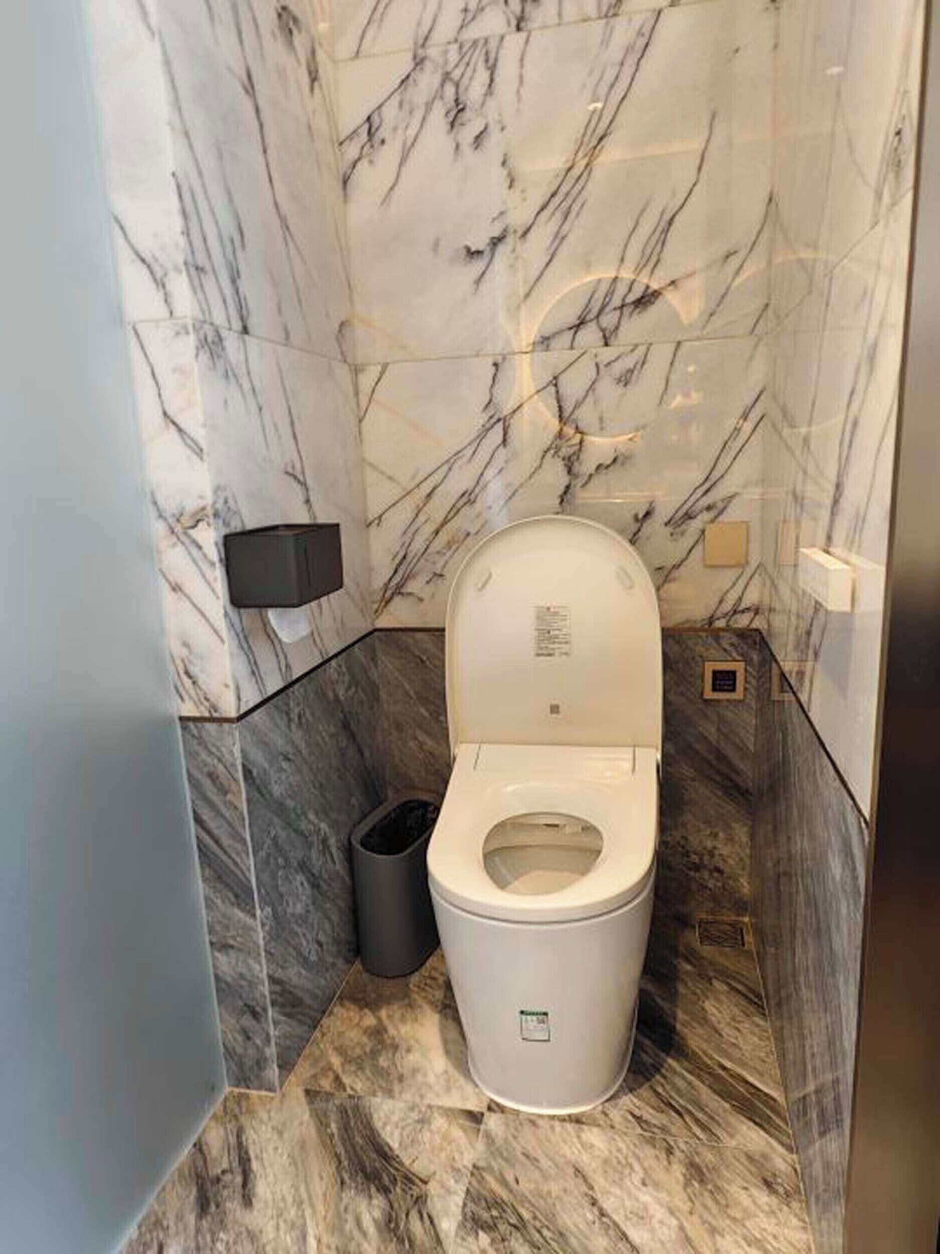 A modern bathroom with a marble floor and walls, a white toilet, and a black trash bin.